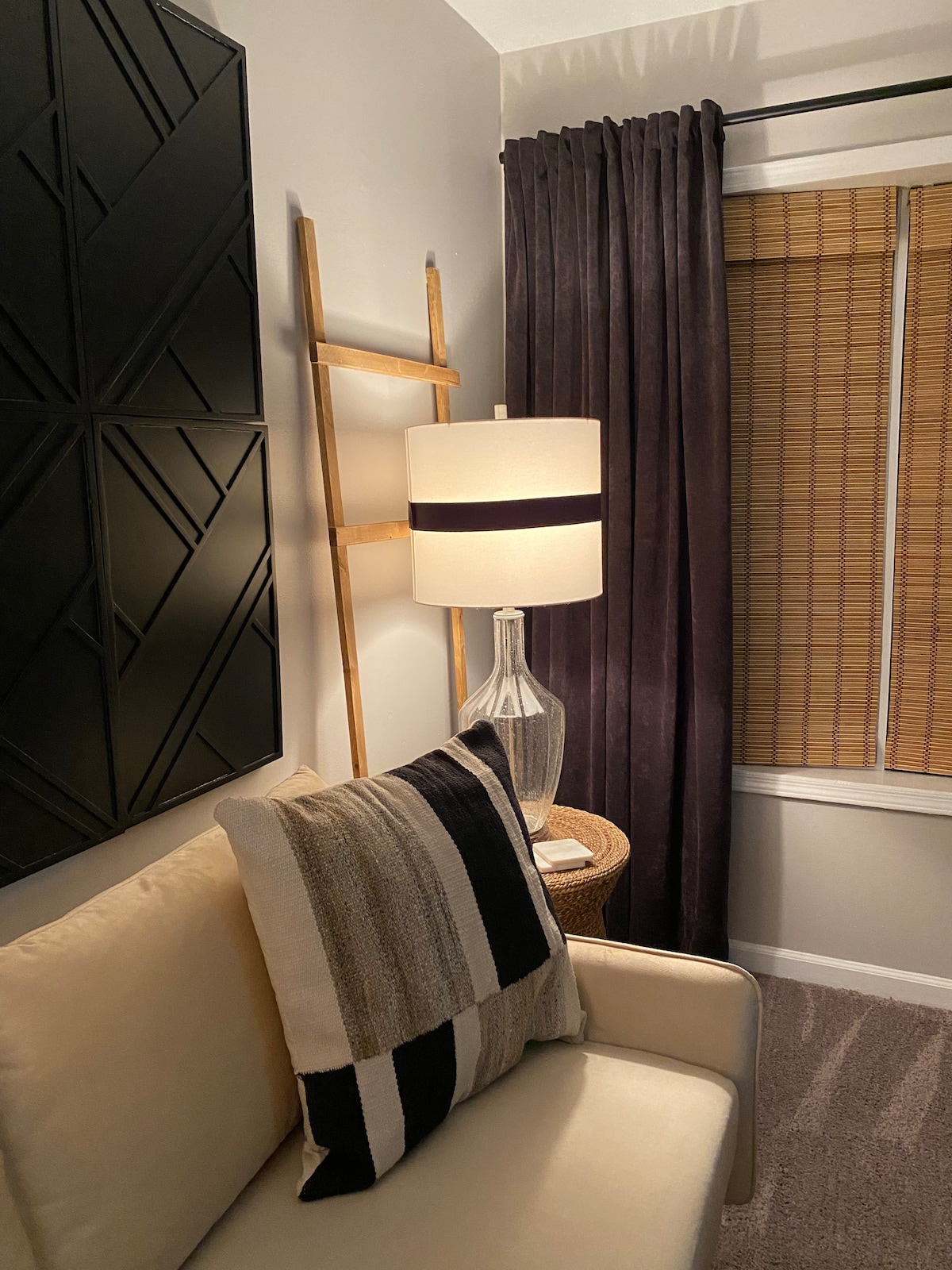 A cozy seating area features a light-colored sofa adorned with a striped cushion. Beside it, a sleek floor lamp with a contrasting shade illuminates the space. Dark curtains frame the window, while a wooden ladder-style decor piece leans against the wall, adding texture.
