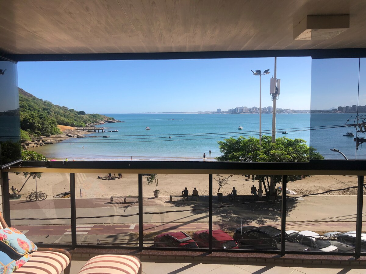 A scenic view from the balcony displays calm waters and sandy beach, with gentle waves lapping at the shore. The coastline is flanked by lush greenery and small boats dot the water. People can be seen enjoying the beach and promenade below.