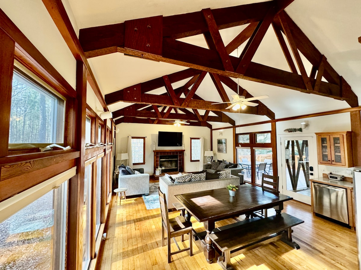 An open living area features high wooden beams and two ceiling fans. A large dining table is centrally positioned, surrounded by natural light from windows. A stone fireplace serves as a focal point, and a cozy seating arrangement is visible in the background.