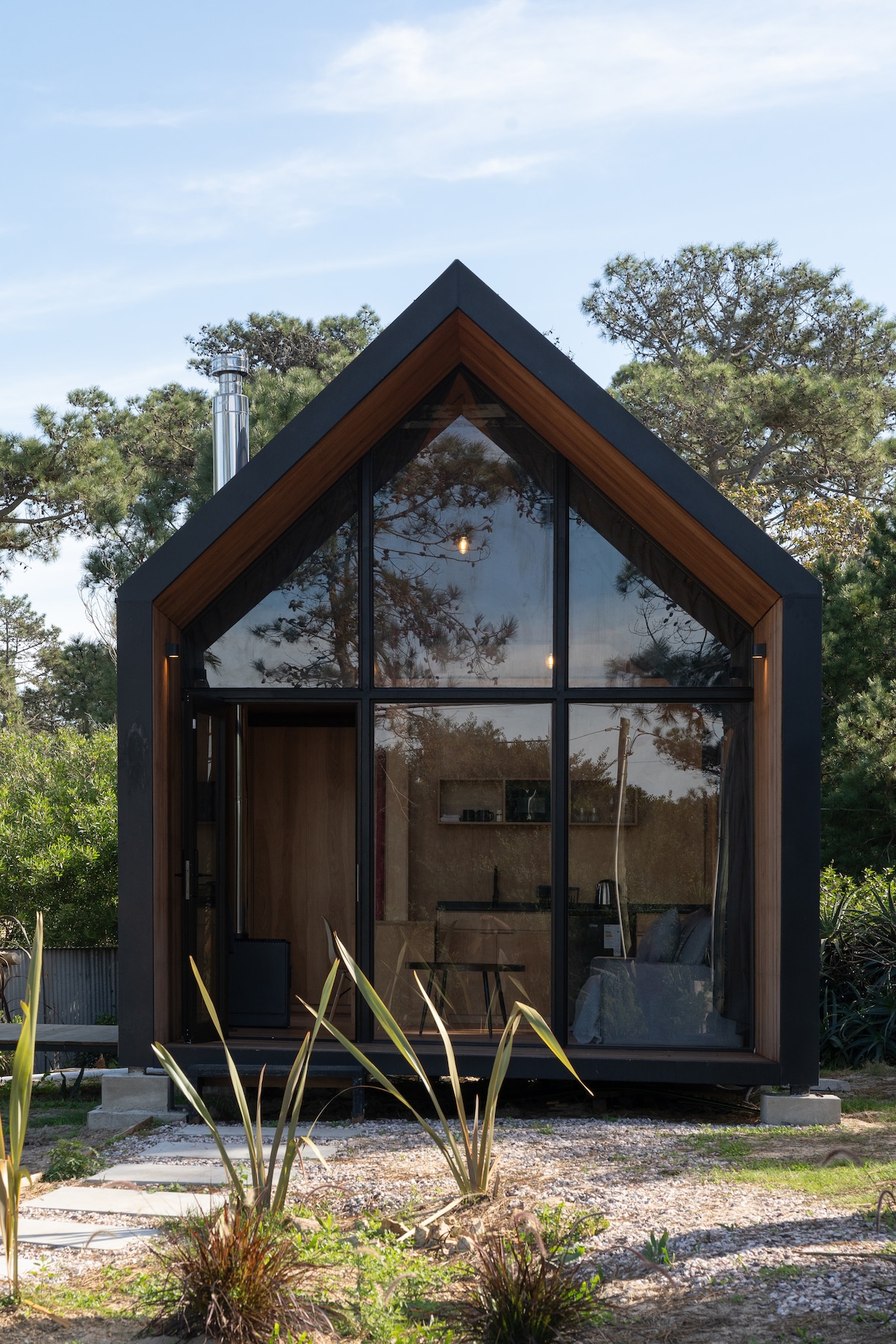 A modern cabin with an angular roof is displayed, featuring large glass panels that bring the outdoors in. The entrance is framed by minimal landscaping, highlighting natural elements. A chimney rises from the rooftop, while the interior is faintly visible through the glass.