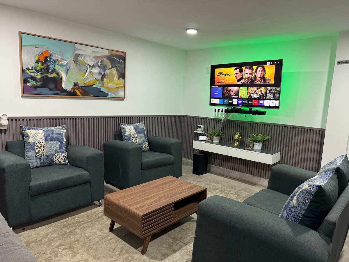 A welcoming living area showcases modern furnishings in muted tones. Two comfortable armchairs and a matching sofa surround a low wooden coffee table. A large wall-mounted screen displays streaming options, complemented by decorative artwork above. Soft lighting adds a relaxing ambiance to the space.
