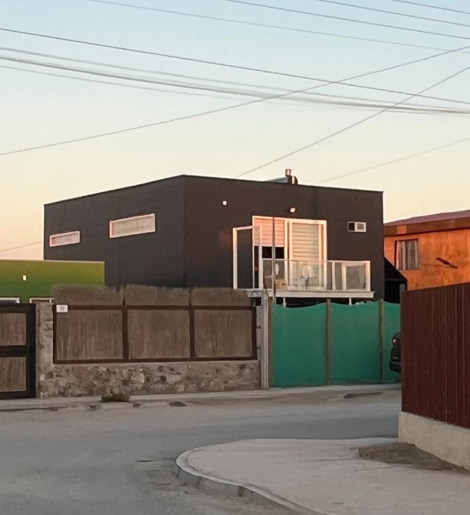 A modern cabin is shown from the street, featuring a dark exterior and large windows. A balcony extends from the upper level, offering a view of the surroundings. A stone wall and a green gate provide privacy, while the road and neighboring structures create a quaint neighborhood atmosphere.