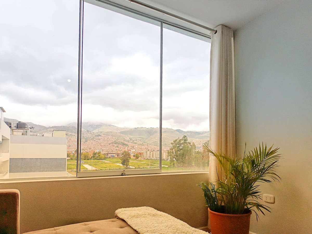 A large window offers an open view of lush green hills and distant mountains. A potted plant resides beside a cozy bed, adding a touch of nature to the indoor space. Soft natural light enhances the calming atmosphere.