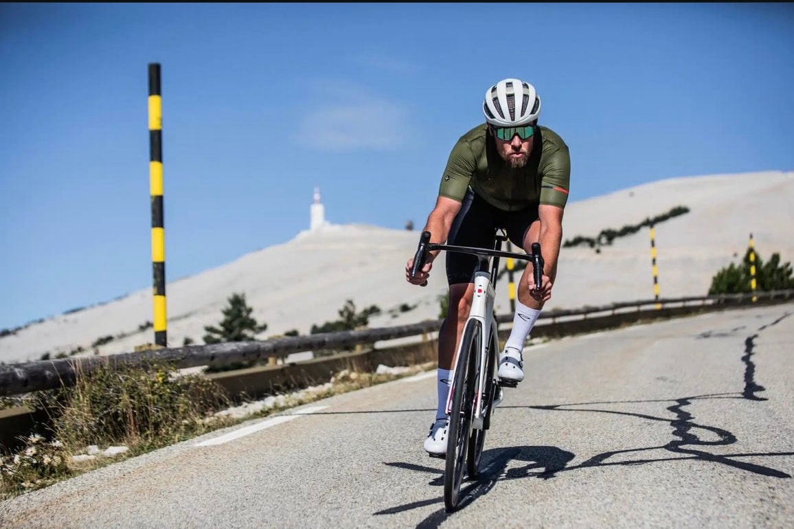 A cyclist is seen riding on a winding road with a backdrop of a prominent mountain. The white rocky terrain of the mountain contrasts with the clear blue sky. The cyclist is dressed in athletic attire and helmet, focused on the path ahead.