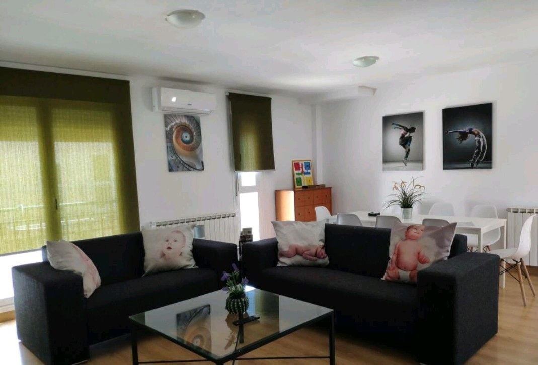 The living area is equipped with two dark sofas adorned with decorative pillows. A glass coffee table sits at the center, accompanied by a small floral arrangement. Natural light fills the space through large windows, which are covered with green blinds. Art pieces are displayed on the walls.