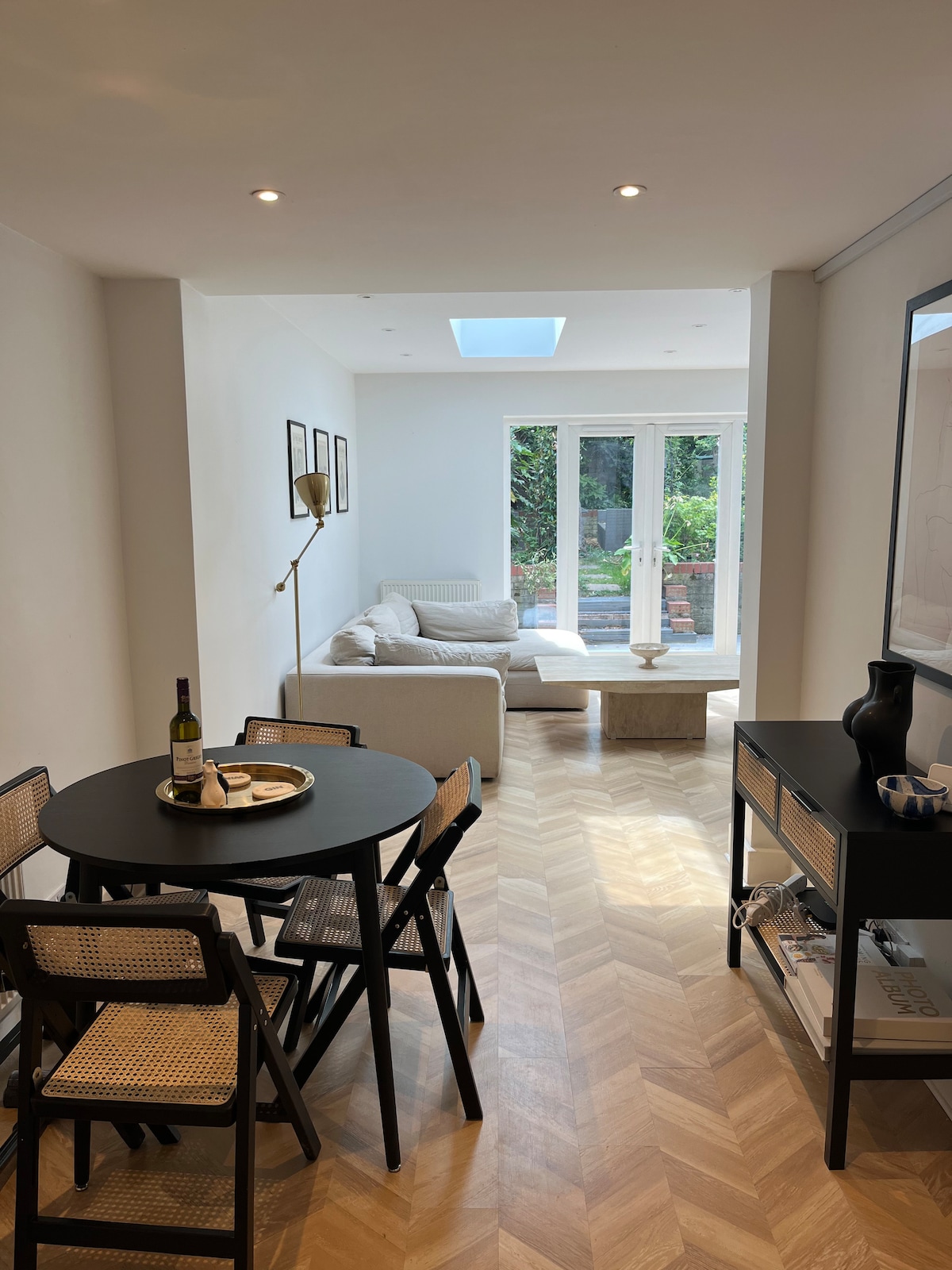 A bright living space is highlighted by a black dining table set for four and a cozy seating area with a light-colored sofa. Skylights provide natural light, and large glass doors open to a private garden, emphasizing the spaciousness of the flat.