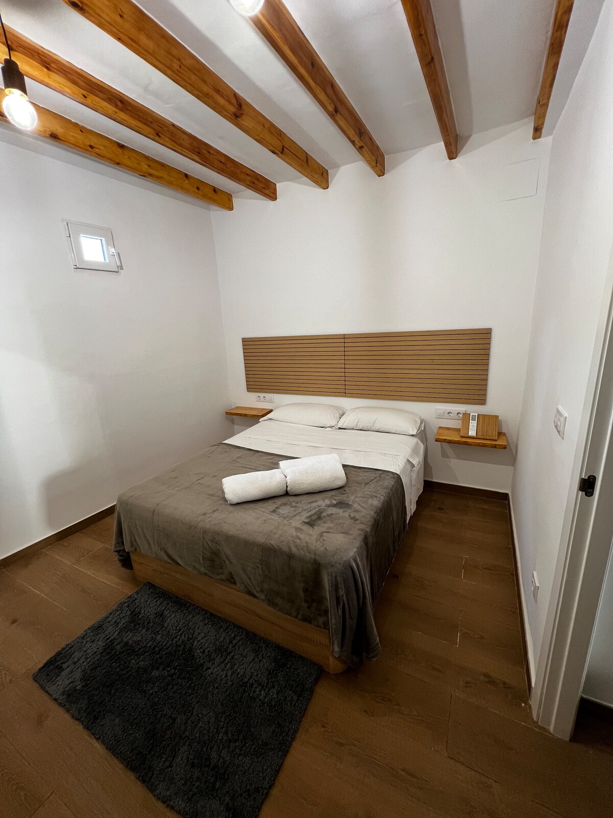 A sleeping area features wooden ceiling beams and soft white walls. The spacious bed is neatly made with crisp linen and two rolled towels. Soft, textured flooring complements the room's design, while natural light enters through a small window.