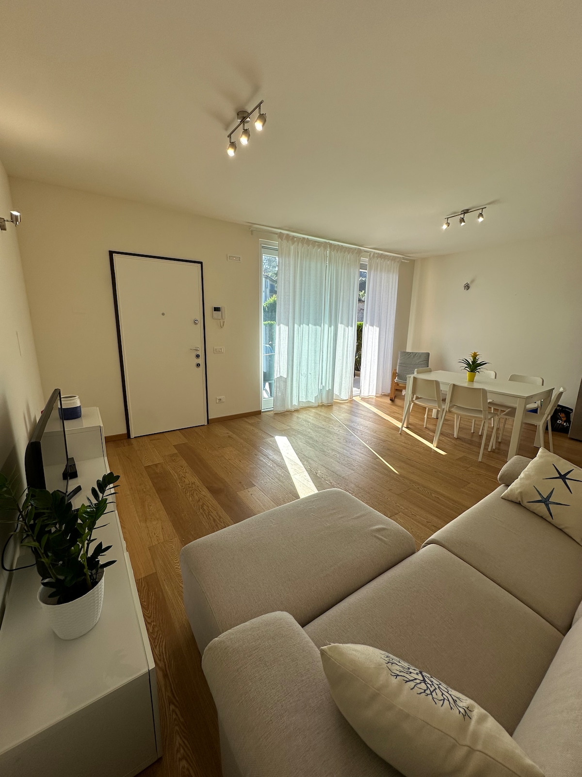 A bright living area features a light-colored sofa and a sleek television stand. Natural light floods through large windows covered with sheer curtains, illuminating the dining table set for four. A small potted plant adds a touch of greenery to the space.