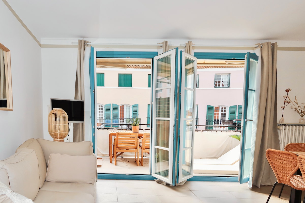 French doors open to a furnished terrace, offering a view of neighboring buildings with green shutters. A light-colored sofa and rattan chairs create a cozy living space. The area is filled with natural light, enhancing the welcoming environment.