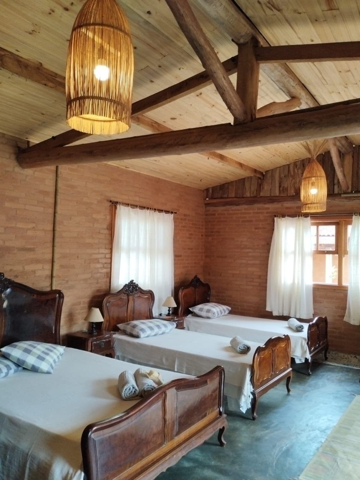 A cozy bedroom features exposed wooden beams in the ceiling, with three wooden beds positioned comfortably in the room. Light-colored bedding is neatly arranged, and decorative pillows add a touch of style. Two large windows allow natural light to brighten the space, complemented by soft curtains.
