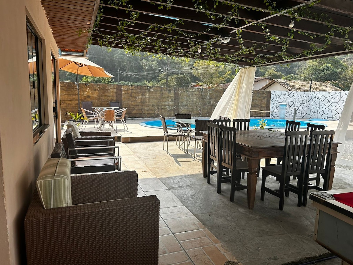 A shaded outdoor seating area features a large wooden dining table surrounded by dining chairs, complemented by a separate lounge area with wicker seating. The nearby pool is visible, framed by paved flooring and surrounded by greenery, creating an inviting space for relaxation.