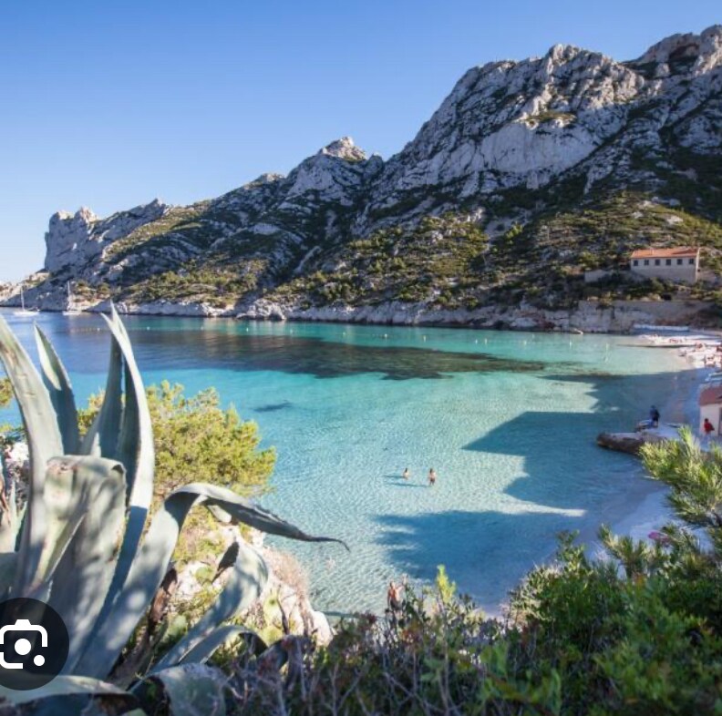 A serene coastal scene is depicted, featuring clear turquoise waters gently lapping at a rocky shore. Lush greenery frames the foreground, while mountains rise majestically in the background. A small beach area is visible, with minimal activity in the water, suggesting a peaceful atmosphere.