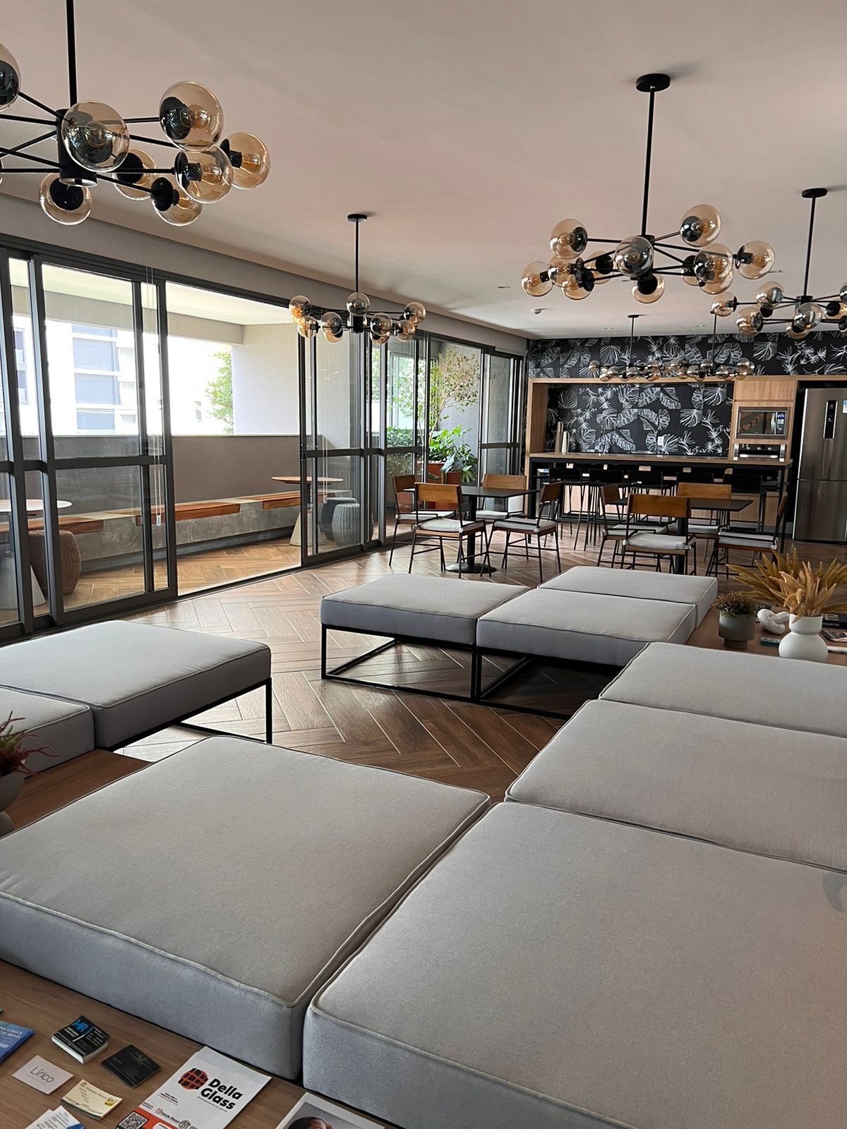 A spacious common area is shown with modern seating arrangements featuring light gray upholstered benches. Large windows allow natural light to fill the room, highlighting a central dining area with wooden tables and chairs. A contemporary kitchen space is visible in the background.