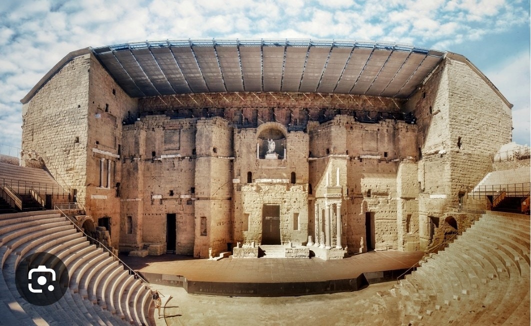 An ancient stone amphitheater showcases impressive architecture with tiered seating arranged in a semicircular layout. The stage area is visible at the center, framed by detailed façades. Skies filled with scattered clouds provide a soft backdrop to the historic structure, emphasizing its grandeur.
