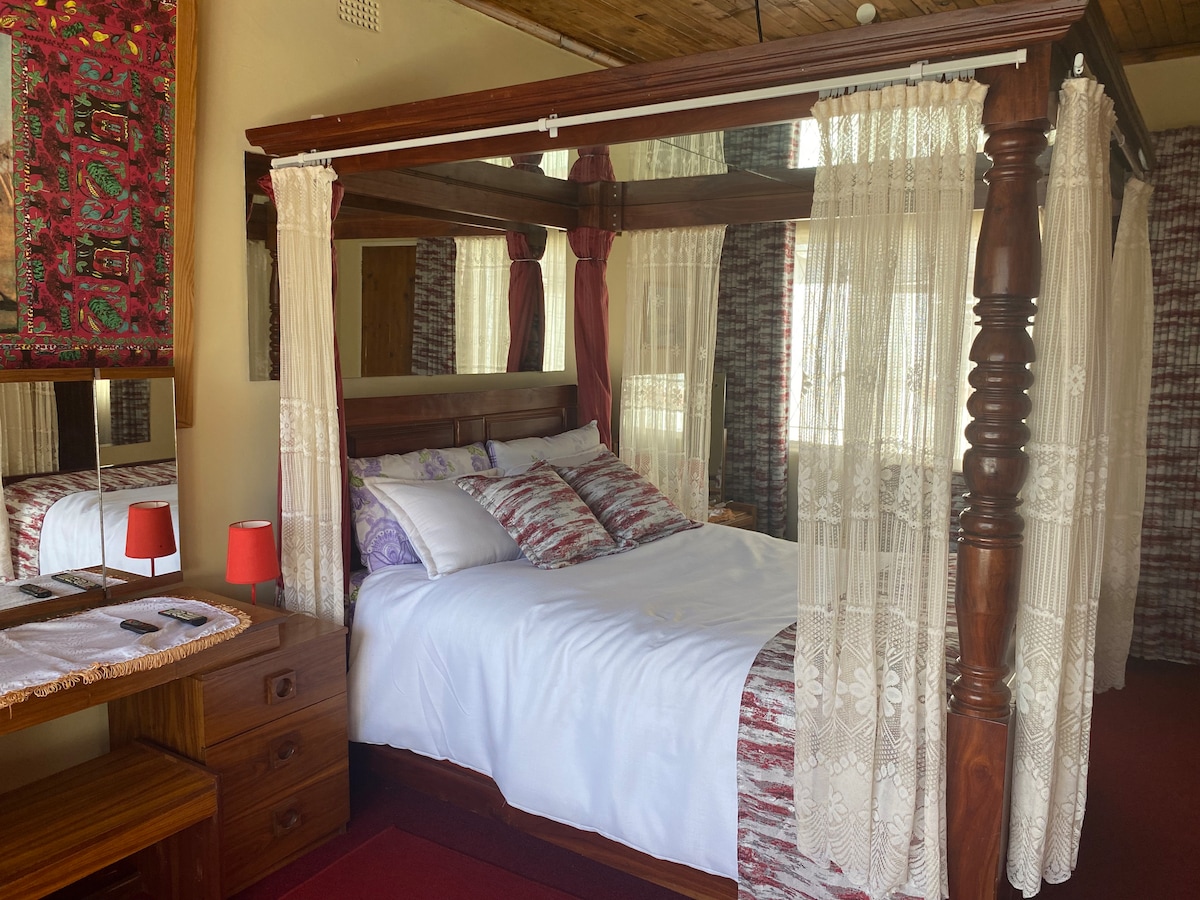 A canopied four-poster bed is elegantly framed by sheer white curtains. The bed is adorned with a decorative red and purple bedspread. A wooden dresser with a vibrant tapestry above it completes the room, which is softly illuminated by natural light entering through a nearby window.