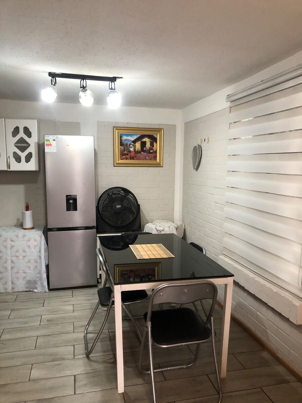 The kitchen area features a modern refrigerator and a black dining table set for four. A fan stands beside the table, while a small decorative piece adorns the wall. Natural light filters through the window, equipped with horizontal blinds.