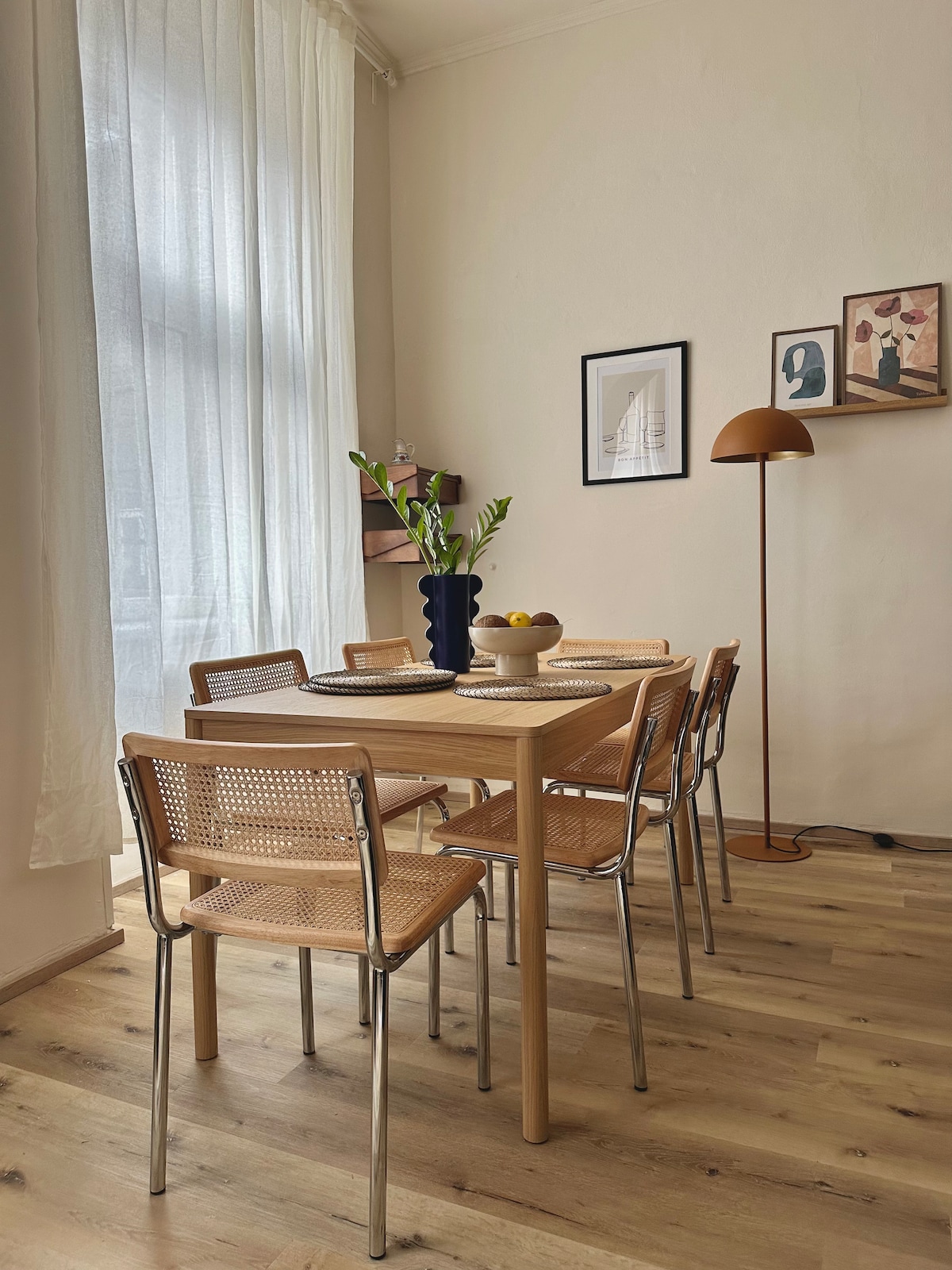 A bright dining area is showcased with a wooden table surrounded by six chairs, featuring a blend of metal and wicker designs. A decorative vase with fresh flowers sits at the table's center, while artworks adorn the walls, complemented by a standing lamp that provides gentle lighting.