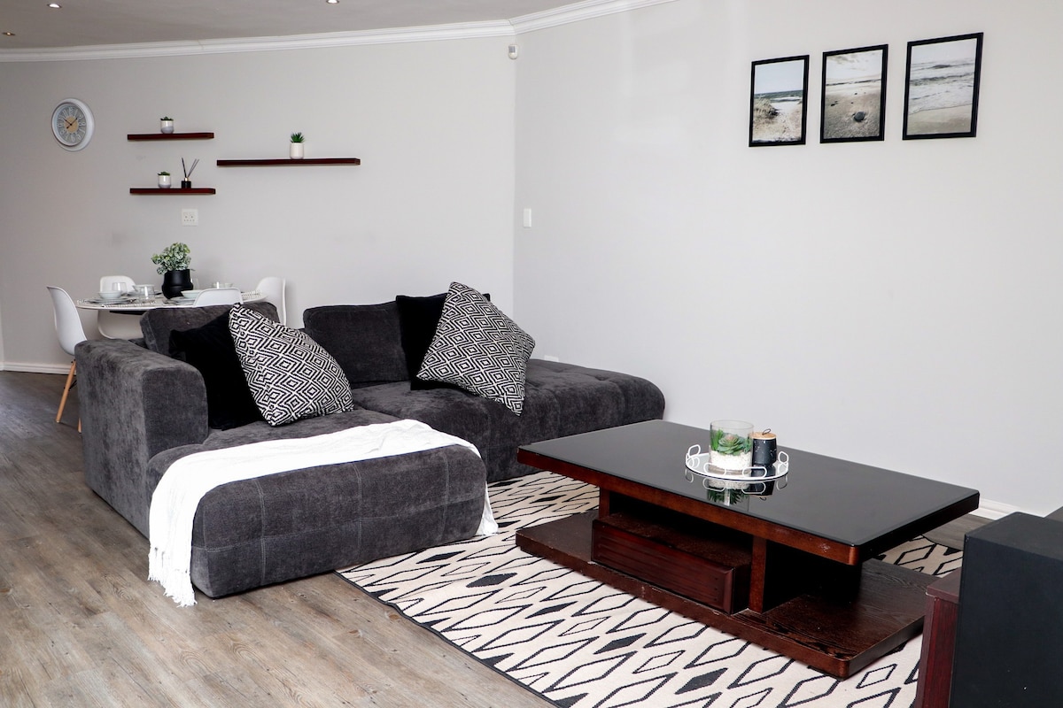 A modern living space features a large, dark sectional sofa adorned with patterned cushions. A low coffee table sits atop a geometric area rug, complemented by simple decor items. In the background, a dining area with a round table and chairs is visible, against a neutral wall.