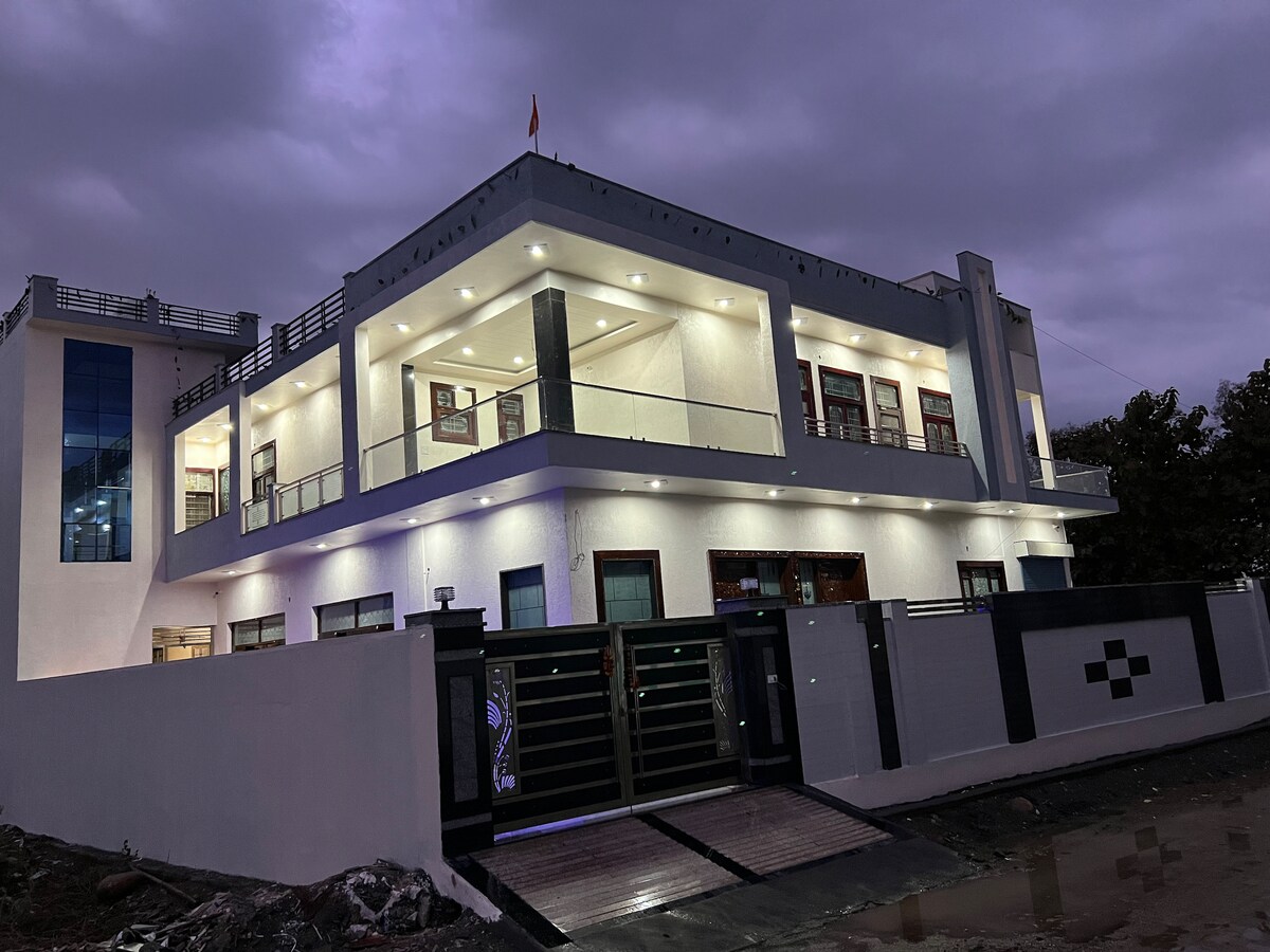 A modern mansion is displayed in twilight, featuring multiple balconies and large windows that illuminate the exterior with warm lighting. The building's clean lines and contemporary design are highlighted against a cloudy sky.