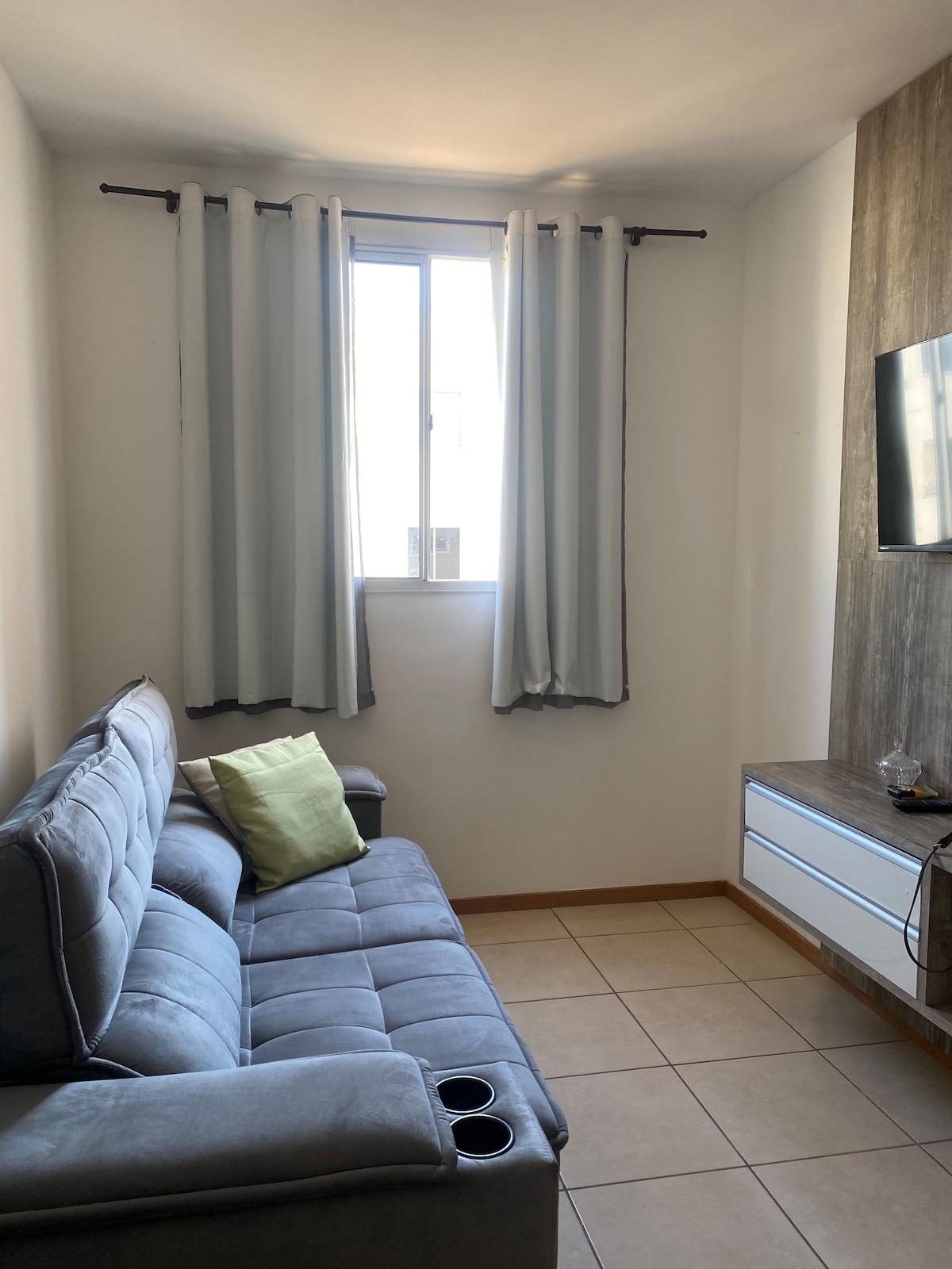 A compact living area features a gray sofa with built-in cup holders. Light curtains frame a window, allowing natural light to illuminate the room. A flat-screen television is mounted on the wall above a small wooden media unit, offering a functional and cozy space.