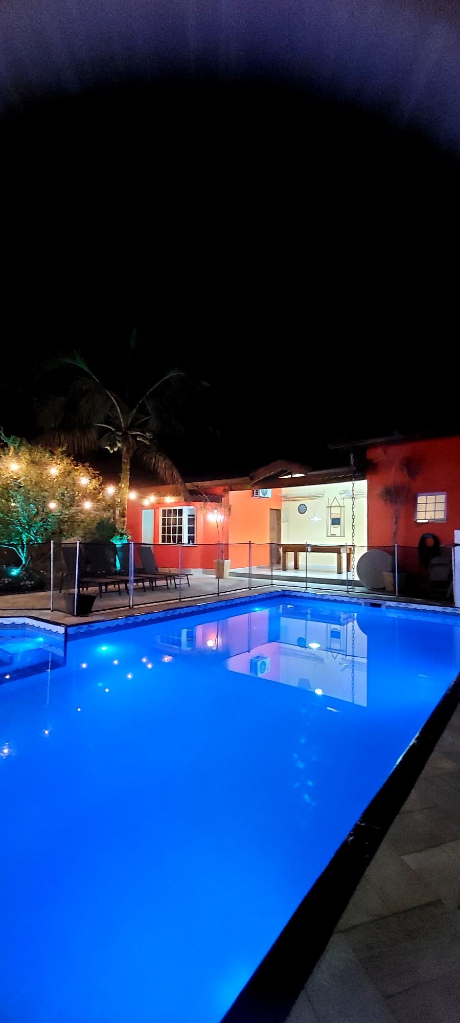 A well-lit pool area showcases a large, illuminated swimming pool reflecting vibrant blue hues. Surrounding greenery is highlighted with soft lighting, adding a touch of warmth to the setting. The house is visible in the background, with welcoming lights and open doors that invite exploration.