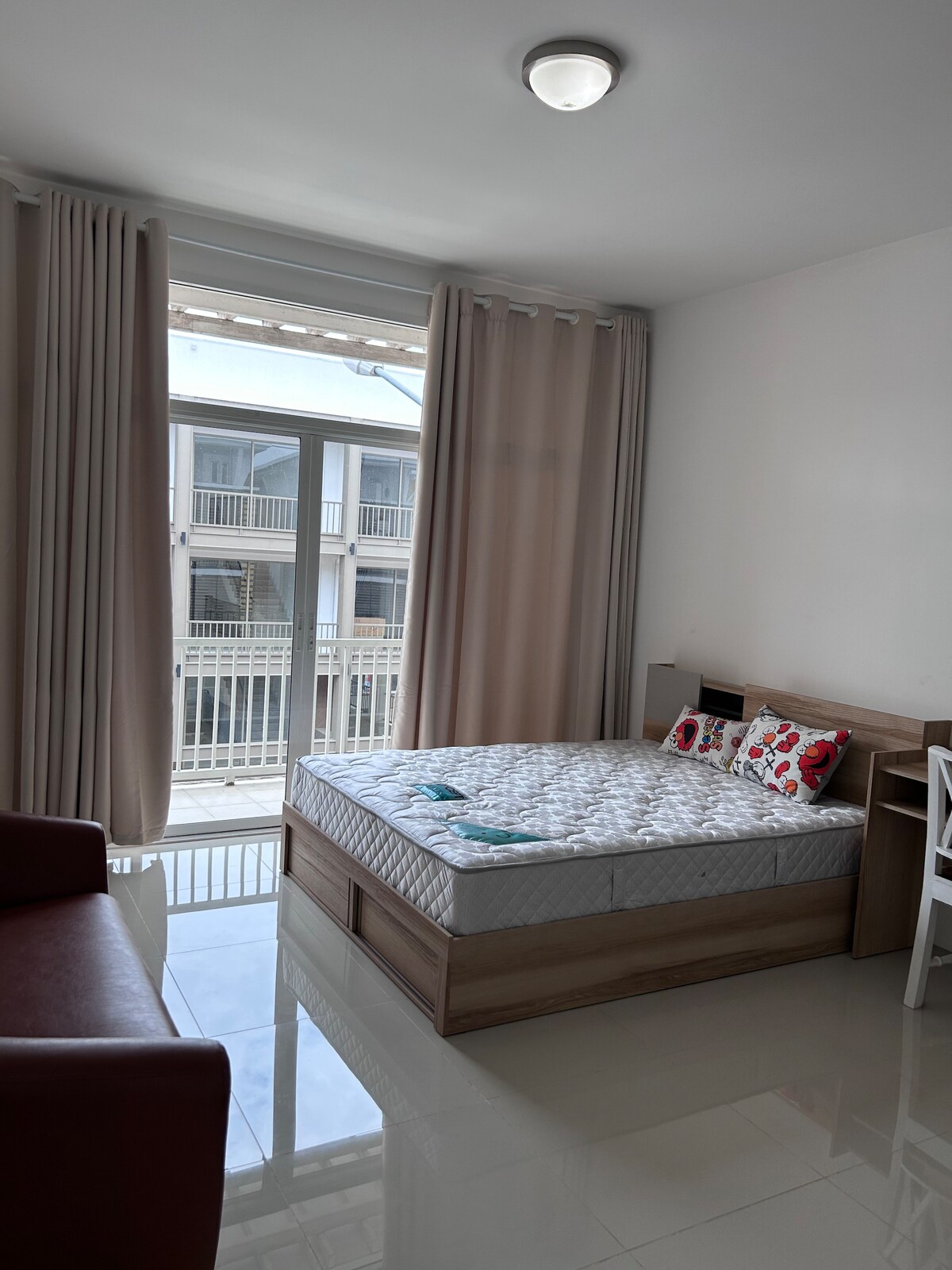 A bright room features a spacious bed made with white linens, complemented by decorative pillows. Large windows allow natural light to fill the space, with sheer curtains gently filtering the sunlight. A small chair is positioned near the bed, enhancing the room's functionality.