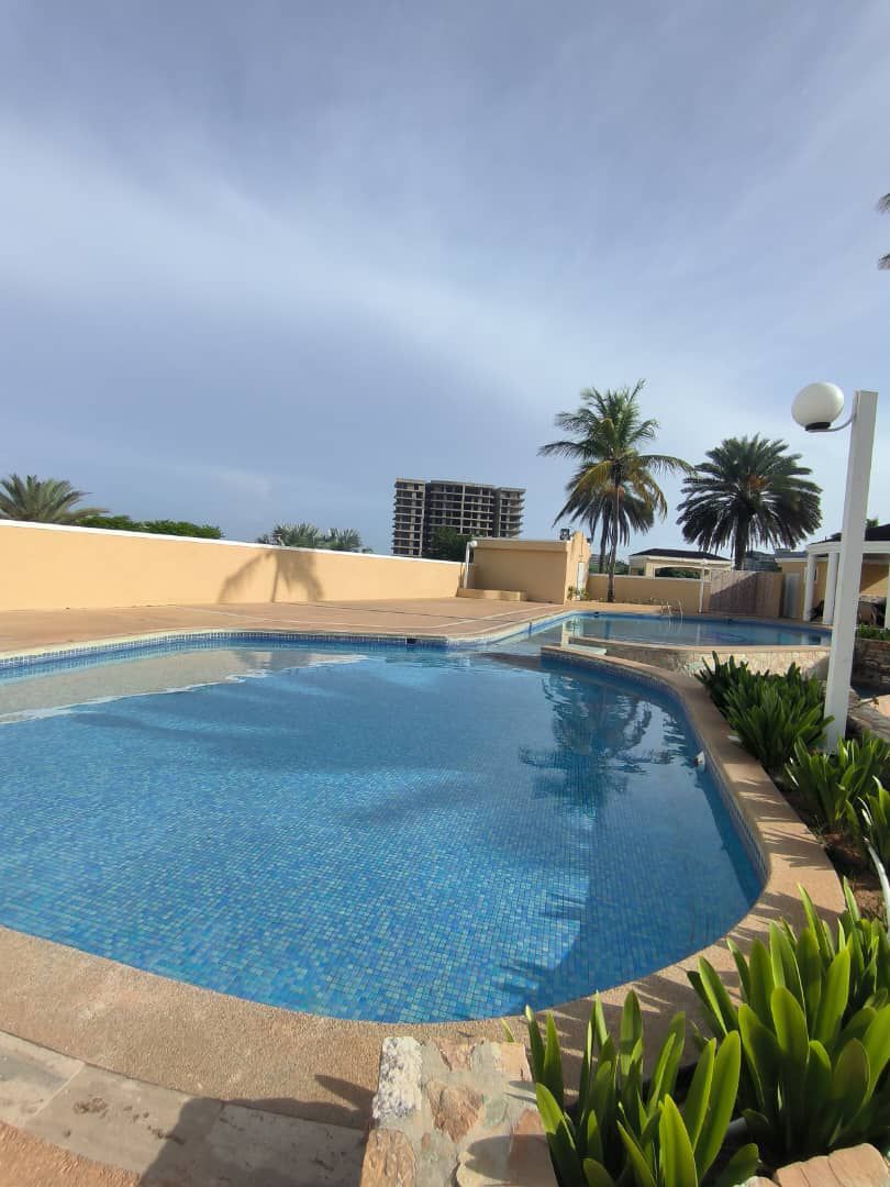 A clear swimming pool reflects the blue sky, surrounded by a patio area with well-maintained green plants and palm trees. The pool's mosaic tiles are visible, adding a tranquil touch to the outdoor space.