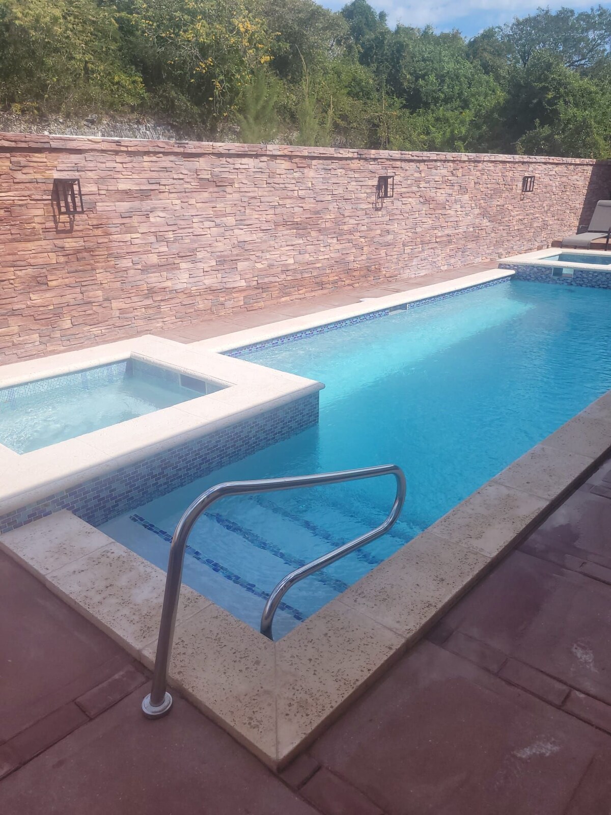 A serene pool area features a spacious rectangular swimming pool with a spa attached. The pool is bordered by a stone wall, and the clear turquoise water reflects sunlight. Sturdy railings offer access to the pool, creating an inviting ambiance for relaxation.