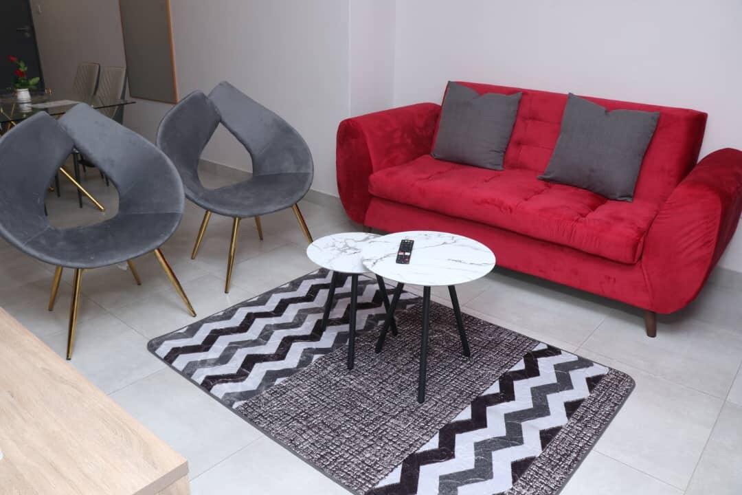 A modern living area is depicted, featuring a red sofa with cushions and two stylish gray chairs. A round marble-top coffee table sits atop a patterned area rug, adding contrast to the neutral tiled floor.