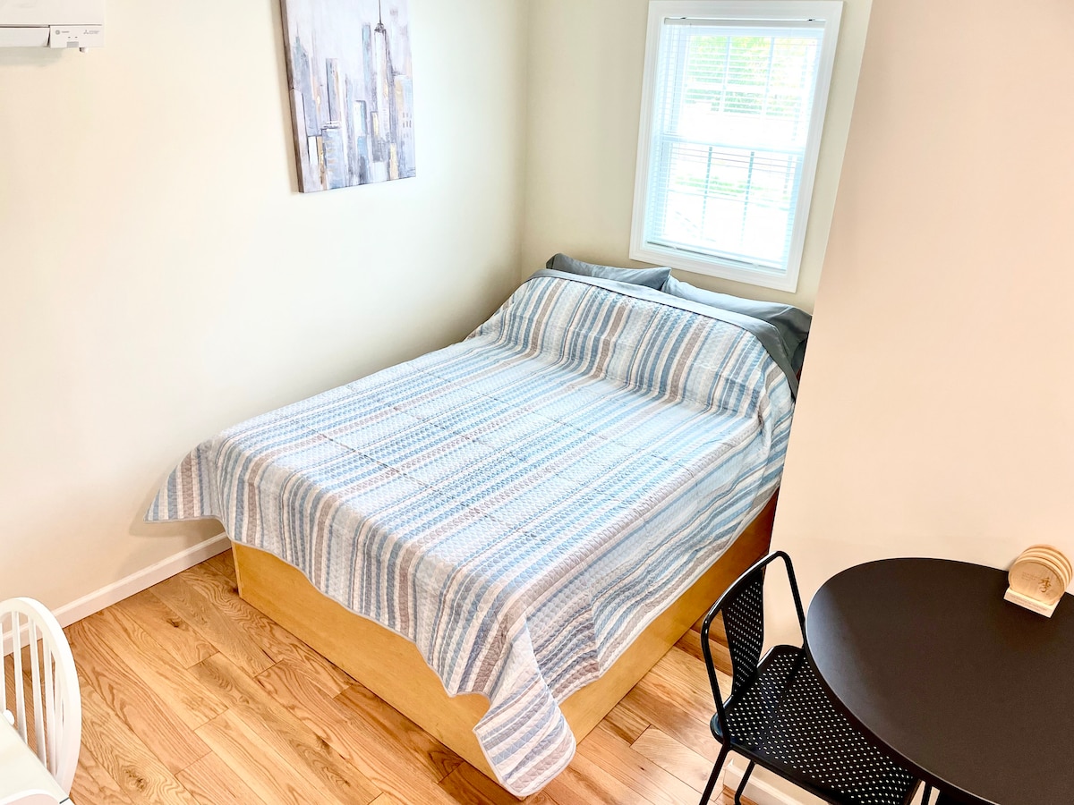A bedroom is depicted featuring a spacious bed covered with a striped blanket. Light enters through a window adorned with simple blinds. A small round table and chairs are positioned nearby, enhancing the functional layout of the space.