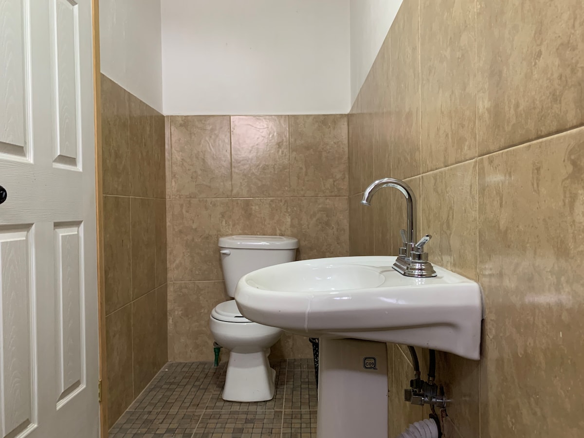 A clean and functional bathroom features beige tiled walls, with a toilet and a sink positioned against one side. The sink has a modern faucet, and the floor is finished with small mosaic tiles.