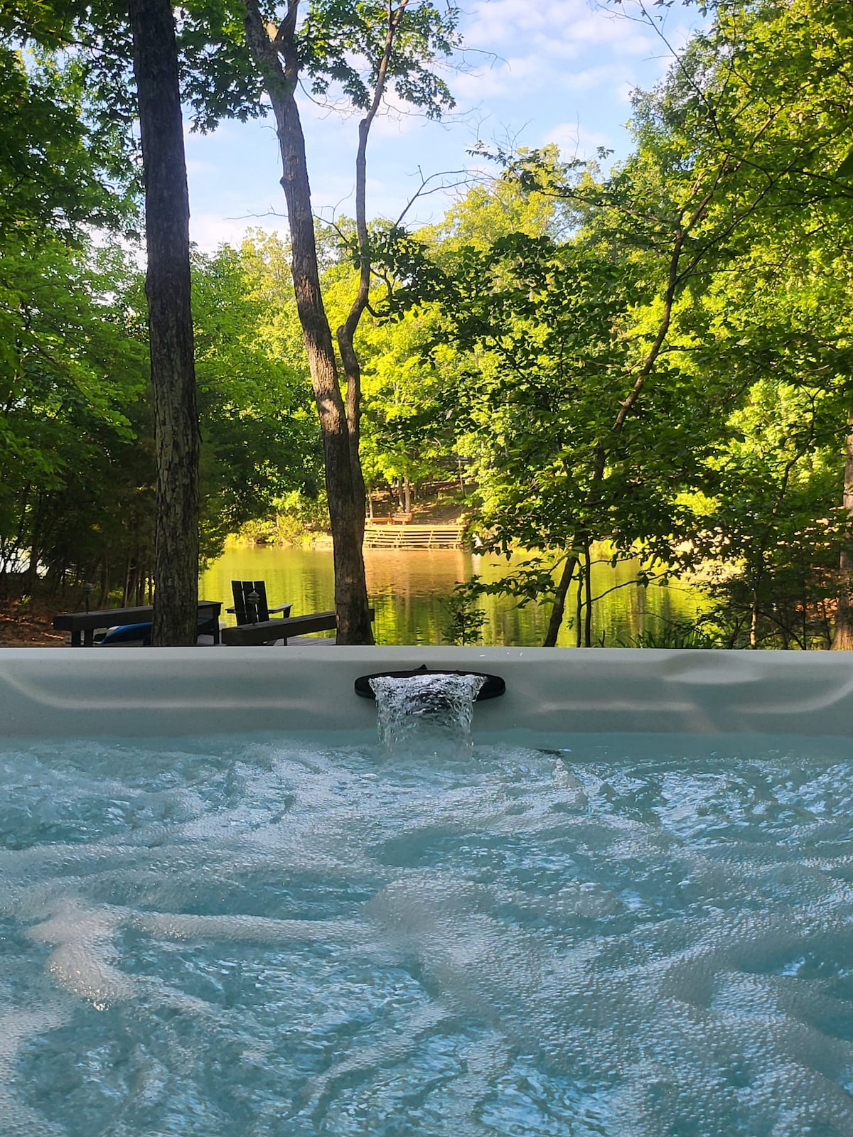 A hot tub is positioned at the edge of the property, with water gently flowing from the spout. Lush green trees surround the area, and a serene lake is visible in the background, creating a peaceful retreat amidst nature.