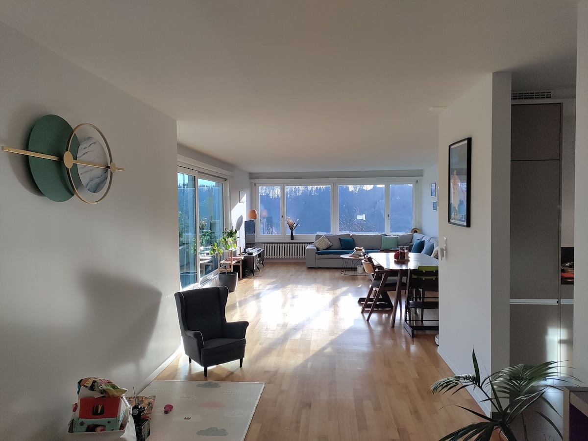 A spacious living area is visible, featuring large windows that allow ample natural light. A dark armchair and a dining table are present, complemented by a sectional sofa. A clock adorns the wall, and potted plants are arranged throughout the space.
