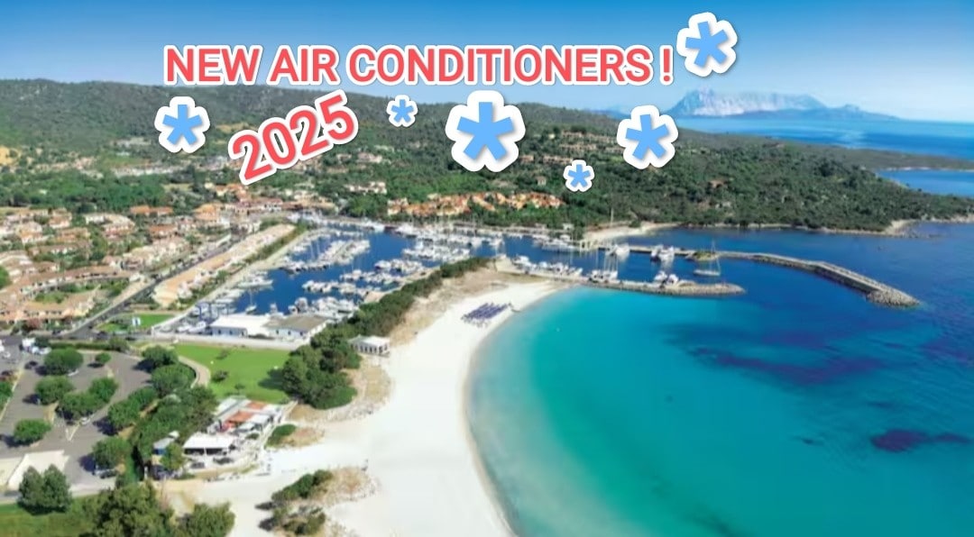 Aerial view of a tranquil beach and marina, showcasing soft white sands meeting clear turquoise waters. Lush green hills frame the scene, and numerous boats populate the marina. The coastline is dotted with trees and structures that provide a serene atmosphere.