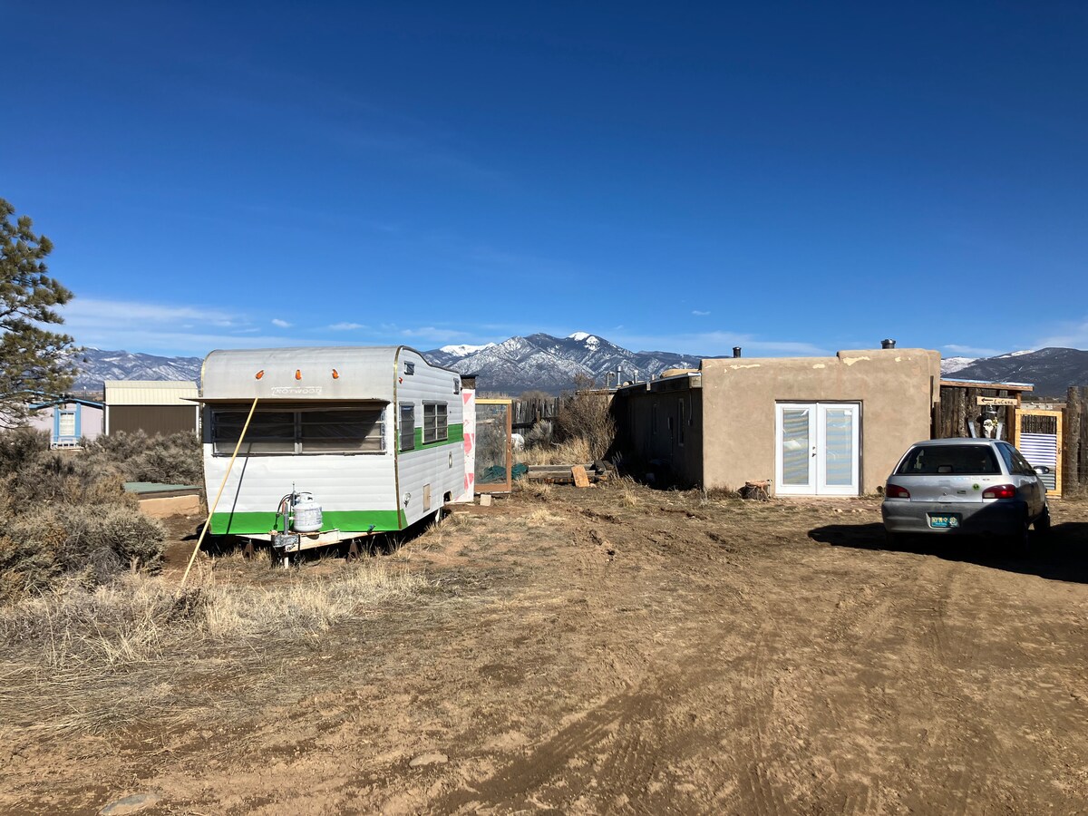 A vintage camper is positioned next to a rustic structure, surrounded by open land with sparse vegetation. The distant mountains are visible under a clear blue sky, contributing to a serene landscape. A parked car can be seen nearby.