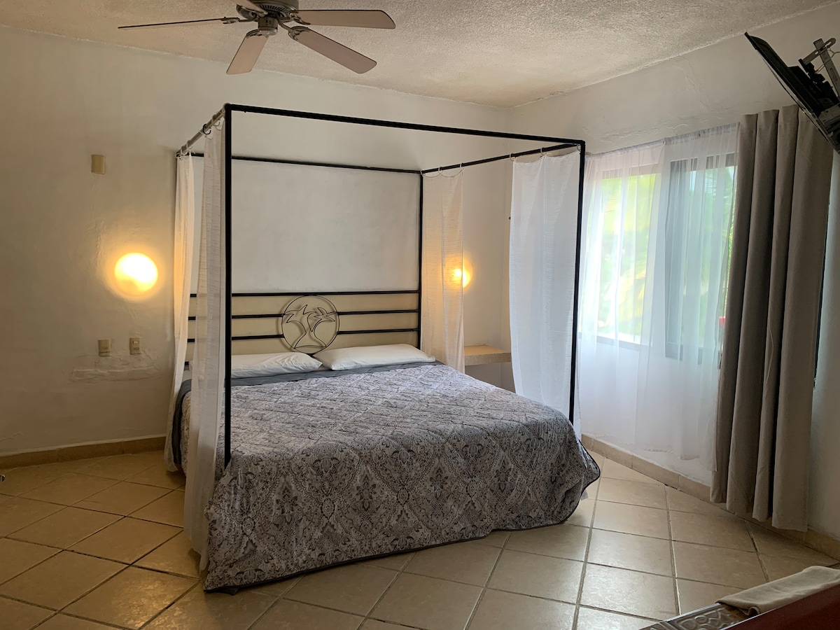 A four-poster bed is showcased in the center of the room, complemented by soothing white drapes. Natural light fills the space through a nearby window, with soft white curtains partially drawn. The tiled floor adds a light and airy feel to the 18 square meter room.