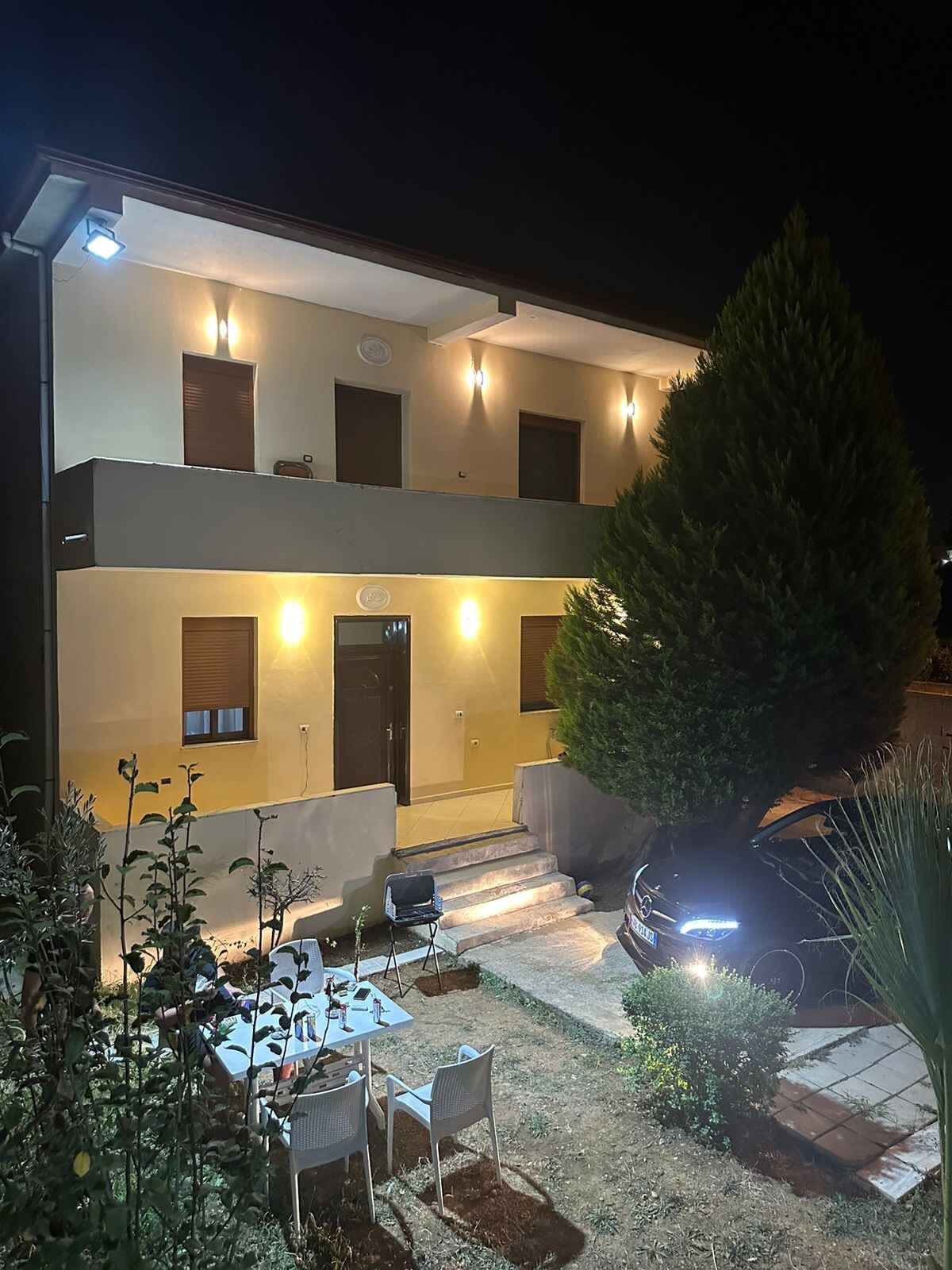 The exterior of a two-story building is illuminated at night, showcasing a welcoming entrance. A set of steps leads up to the door, flanked by well-lit windows. A dining area with a table and chairs is positioned in the foreground, surrounded by greenery.