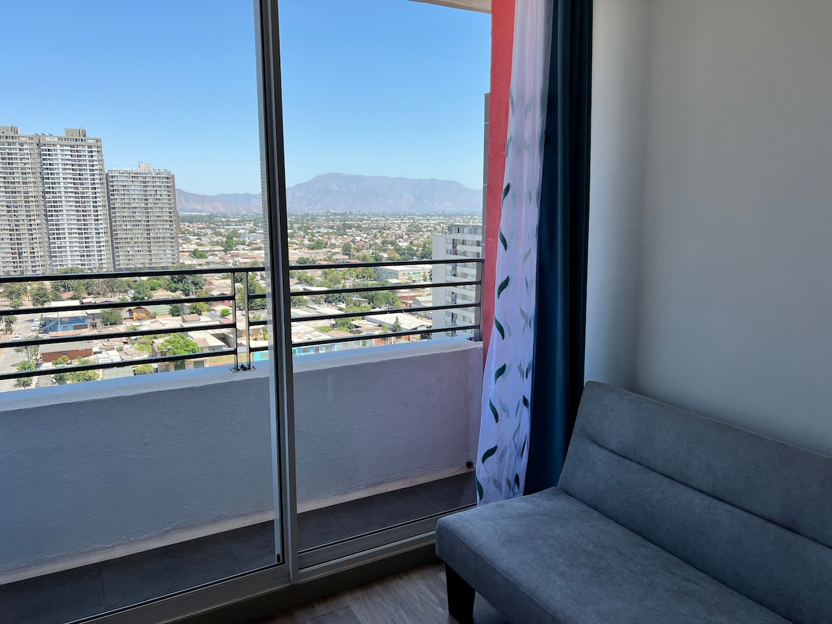 A comfortable seating area is seen with a gray sofa positioned next to a large window, offering expansive views of the cityscape and distant mountains. Natural light fills the space through sheer curtains, enhancing the open feeling.