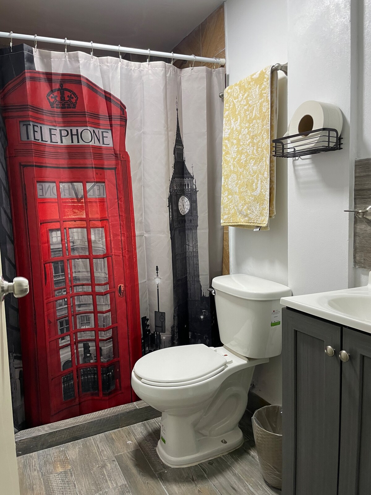 The bathroom features a modern setup, including a white toilet and sink surrounded by gray cabinetry. A unique shower curtain displays a red telephone booth and a clock tower design, while a yellow towel hangs neatly on a rack above.