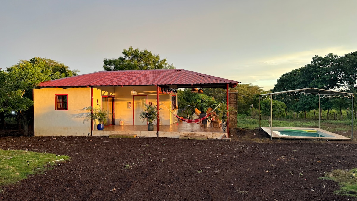 A single-story building features a red roof and light-colored walls, with large front doors flanked by plants. A hammock is visible on the porch, and a shaded outdoor space holds a swimming pool nearby, surrounded by lush greenery.