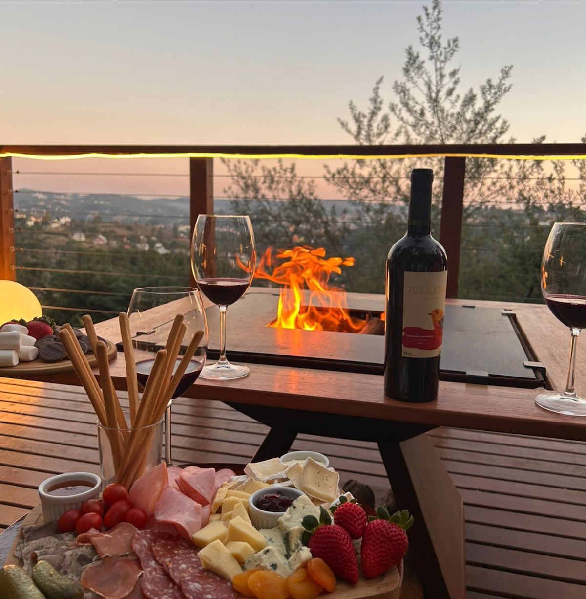 A cozy outdoor dining area features a wooden table set with a charcuterie board, surrounded by glasses of red wine. A small fire pit is visible, adding warmth to the setting. The background showcases a sunset over distant hills, enhancing the serene atmosphere.