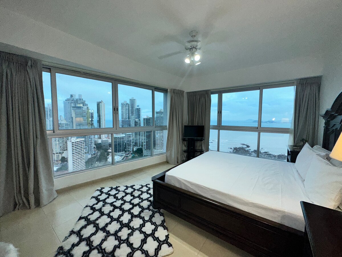 The bedroom features a spacious bed with a dark wood frame. A large window offers panoramic views of the city skyline and bay. Soft curtains frame the window, complementing the light neutral tones of the room. A flat-screen TV is positioned in the corner, enhancing the space.