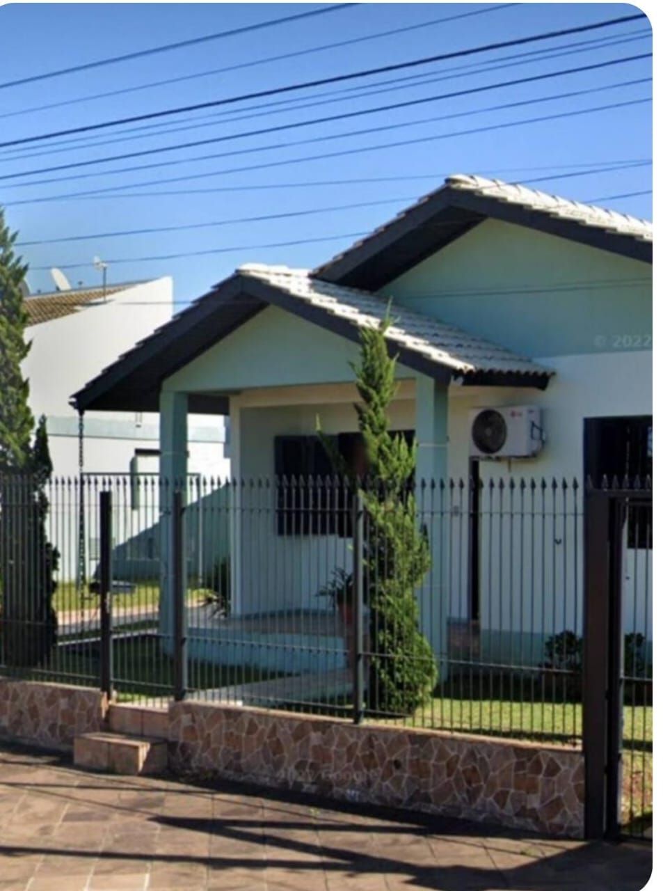 A single-story house is framed by a black wrought iron fence. Two neatly trimmed trees stand in front of the light blue facade, which features a sloped roof and an air conditioning unit. The clear sky overhead enhances the inviting exterior.