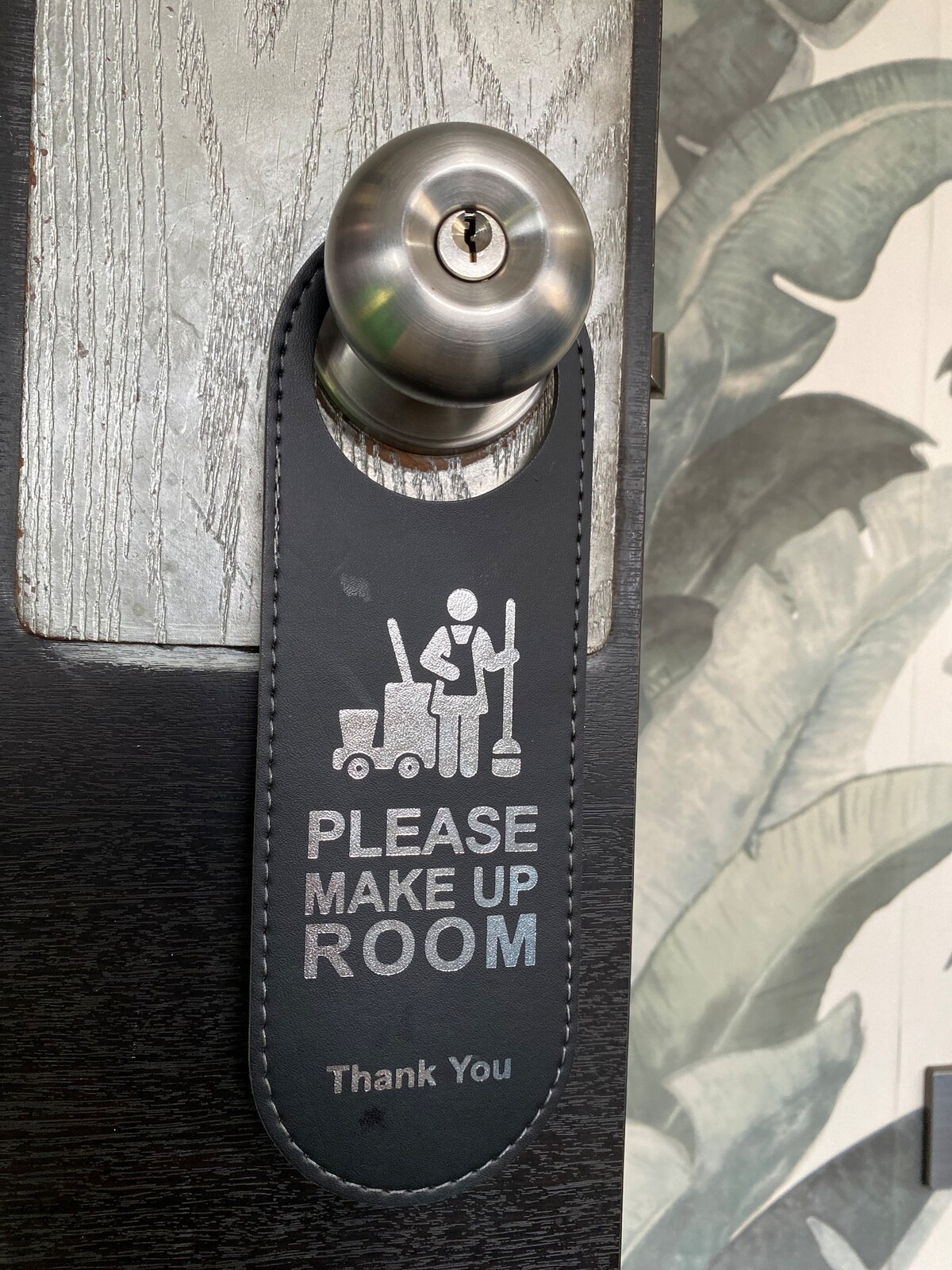 A door sign is displayed on the handle, indicating 'Please Make Up Room.' The sign features a graphic of a person cleaning, emphasizing the request for room maintenance. The door's texture and the backdrop of greenery are subtly visible, enhancing the overall aesthetic.