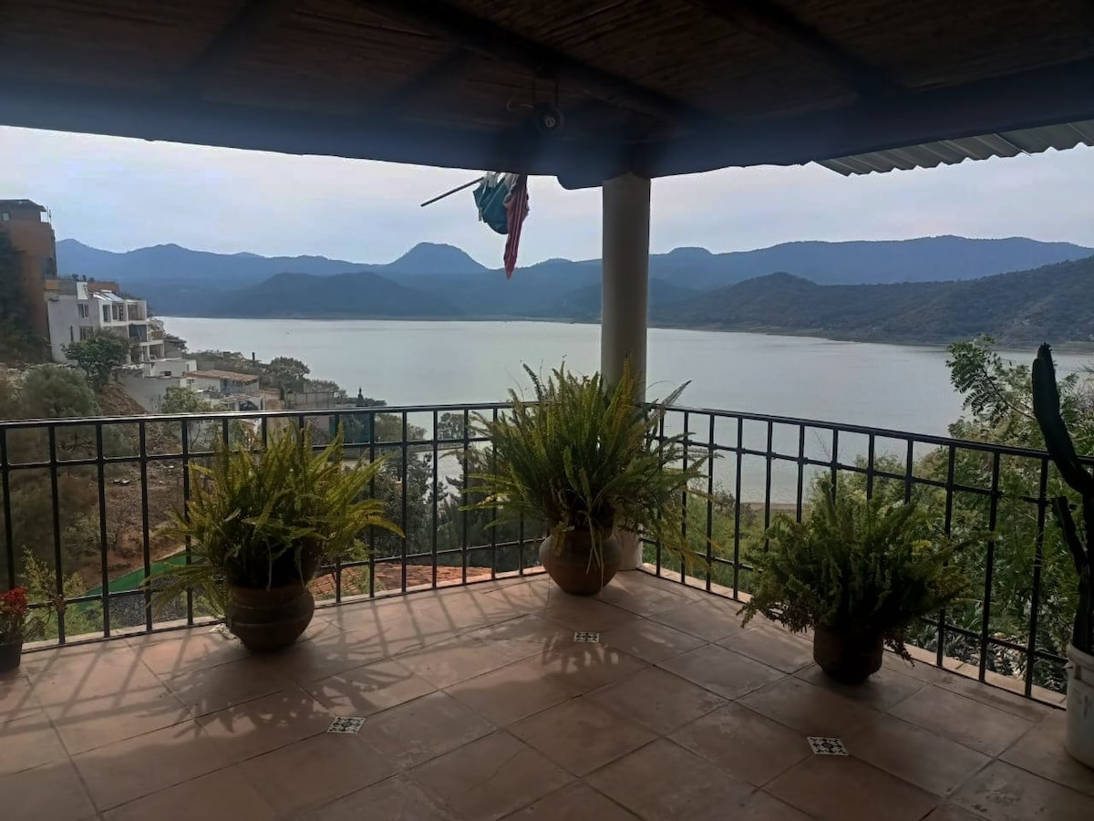 A spacious terrace overlooks a serene lake, framed by lush mountains in the distance. Potted plants add a touch of greenery, while a railing lines the terrace, offering a clear view of the landscape. The sky appears overcast, creating a calm ambience.
