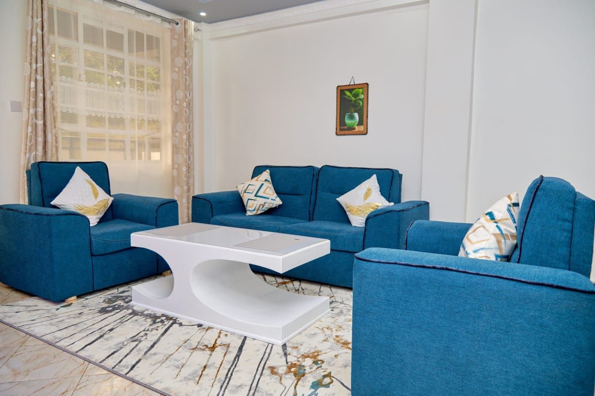 A bright living area features a set of four blue armchairs, each adorned with decorative cushions. A sleek white coffee table occupies the center, contrasting with a light-colored, patterned area rug. Natural light filters in through large windows draped with sheer curtains.