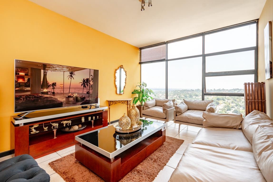 The living area features large windows providing scenic city views. A plush, light-colored couch is arranged around a contemporary coffee table, with a soft area rug beneath. A spacious television stands against the wall, while a decorative mirror and indoor plants add character to the space.