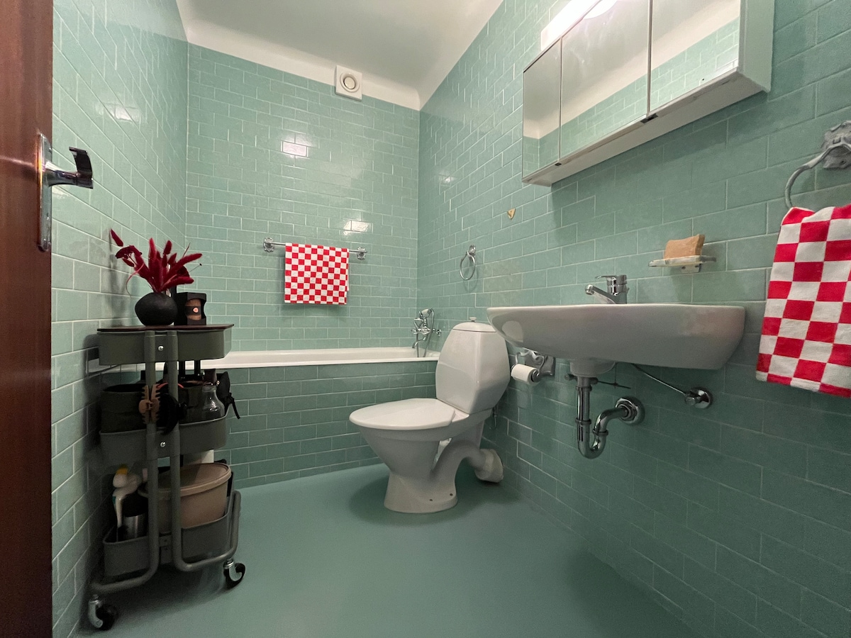 The bathroom features bright teal wall tiles and a clean, modern design. A white toilet is positioned next to a sink, with a mirrored cabinet above. A rolling cart holding toiletries adds convenience, and a red and white checked towel hangs nearby.