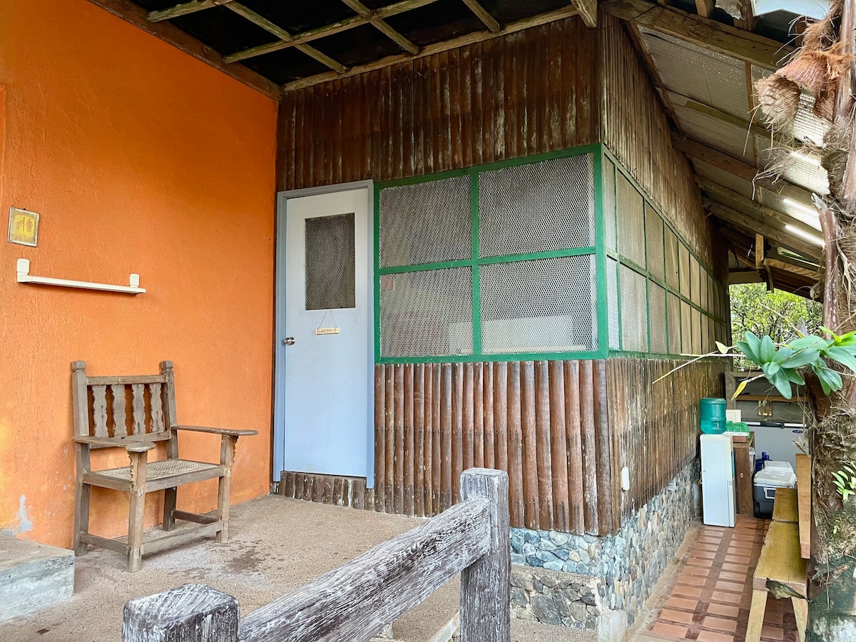 A rustic wooden structure is featured with an orange exterior and a screened porch. A simple wooden bench is positioned in front, providing a space for relaxation. A door leads inside, and an outdoor area with a counter and sink is visible to the side.