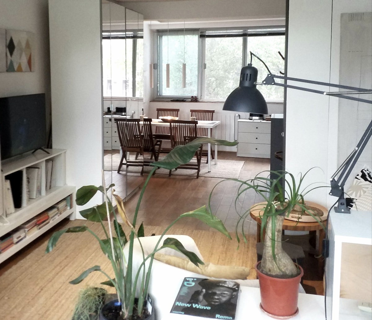 An open concept living area is highlighted, featuring a modern television unit and two green plants. A light-colored dining table surrounded by wooden chairs is visible in the background, with large windows offering views of trees outside. A pendant light hangs above the dining area, enhancing the spacious feel.